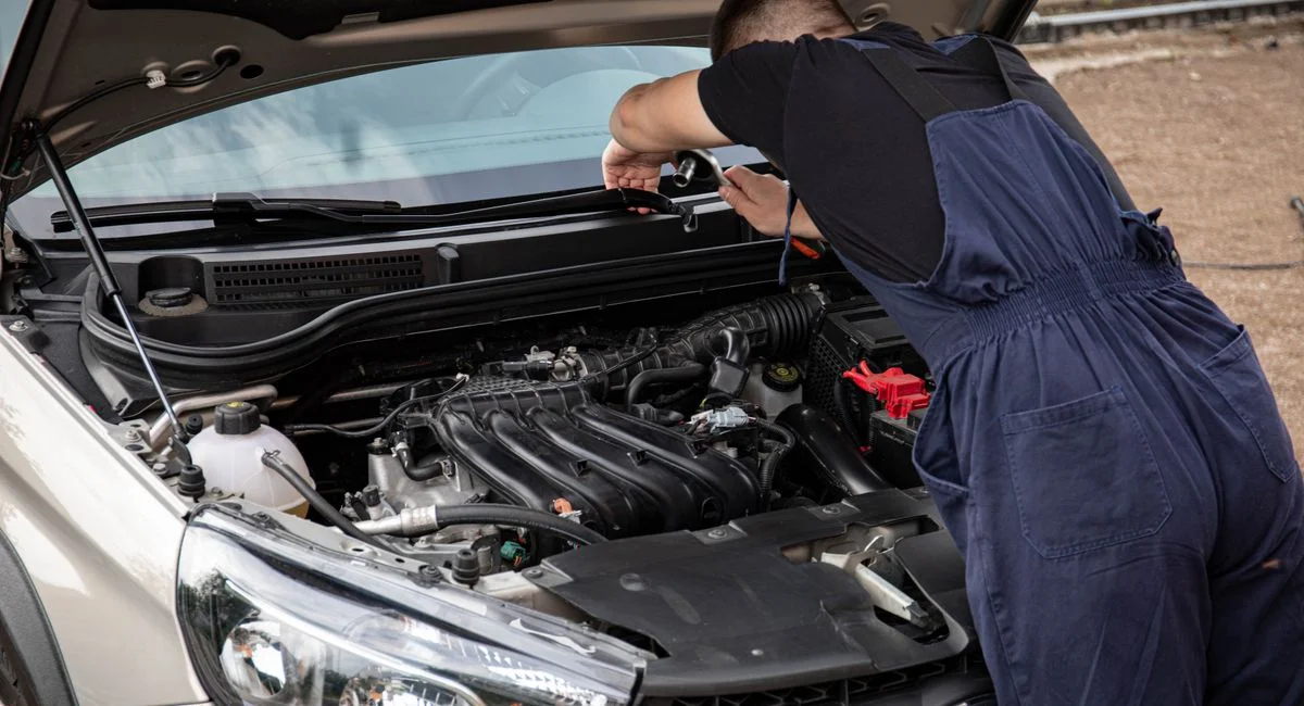 A mechanic in blue overalls works on a car engine in an outdoor setting.