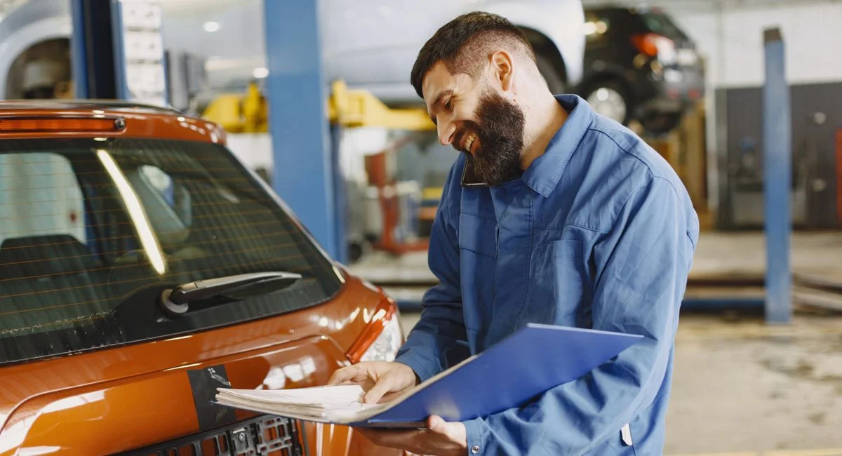 Smiling auto mechanic inspects a car in a service garage, wearing coveralls and holding documents.