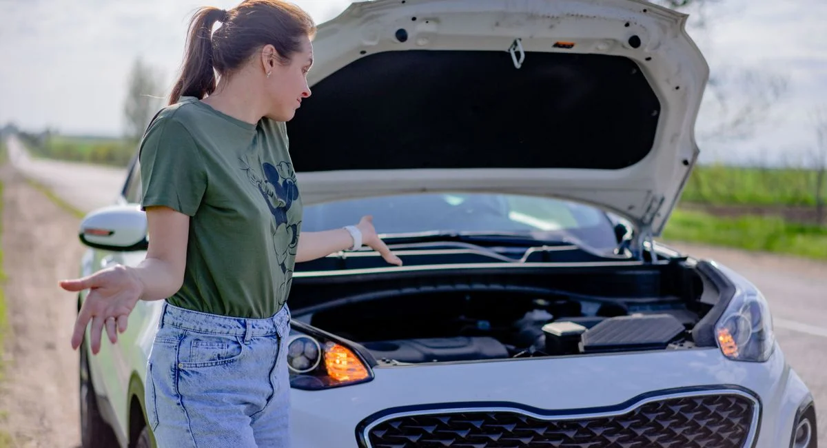 A woman looks puzzled by her broken car with an open hood on a rural road.