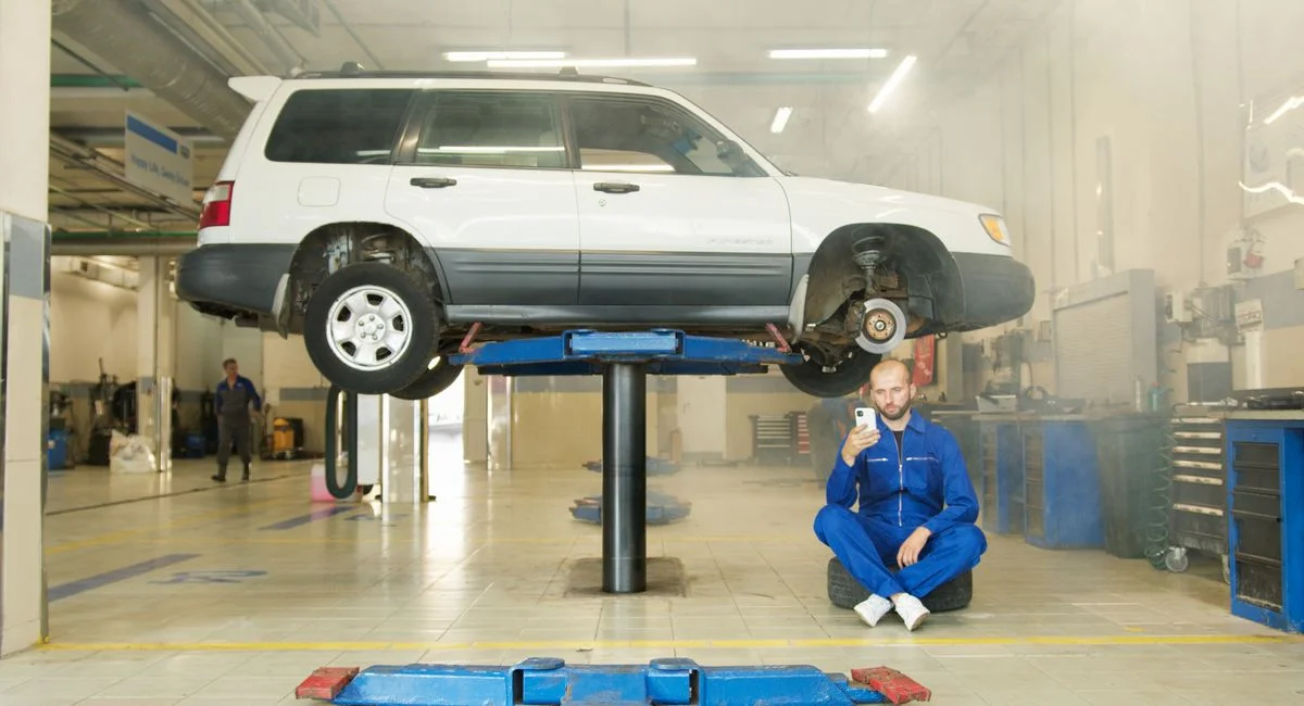 Mechanic in blue coverall using smartphone beside vehicle on lift in repair workshop.