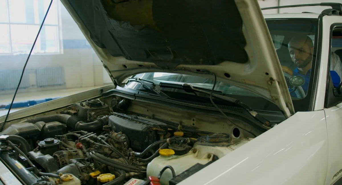 Mechanic checking car engine under open hood in a garage setting.