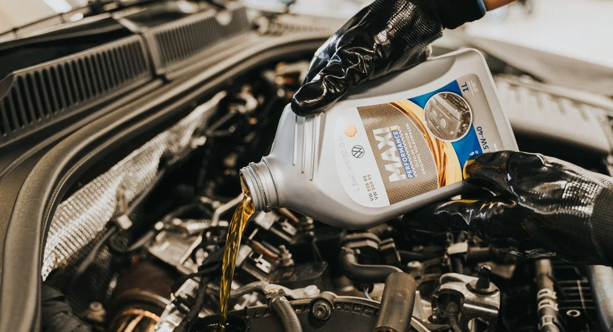 A mechanic wearing gloves pouring oil into a car engine in a garage setting.