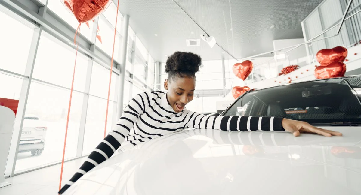 Excited woman embraces her new car at a dealership, surrounded by red balloons.