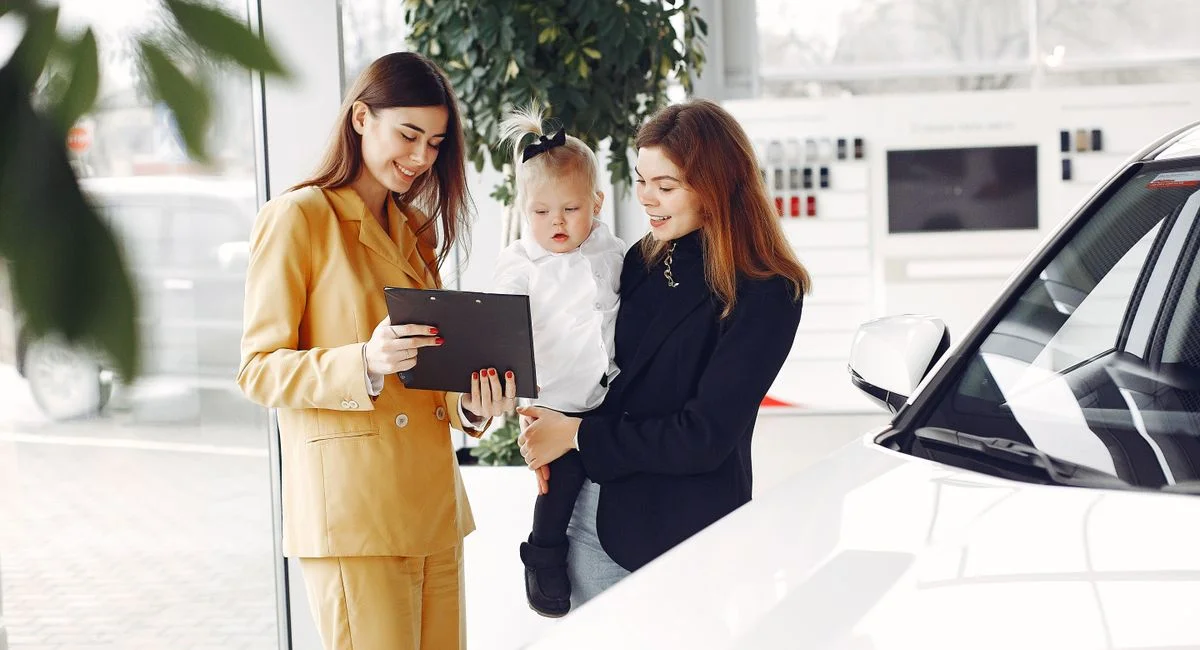 Positive young female with cute little daughter on hands discussing car characteristics with professional dealer in stylish b