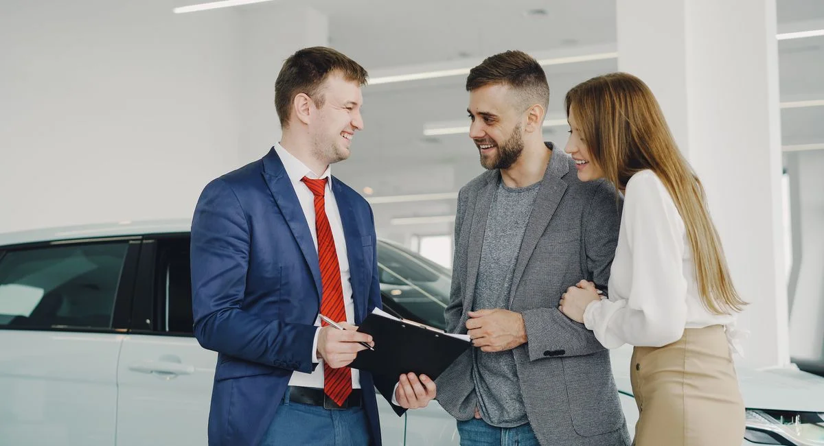 A smiling couple buys a new car from a confident salesman inside a modern car dealership.
