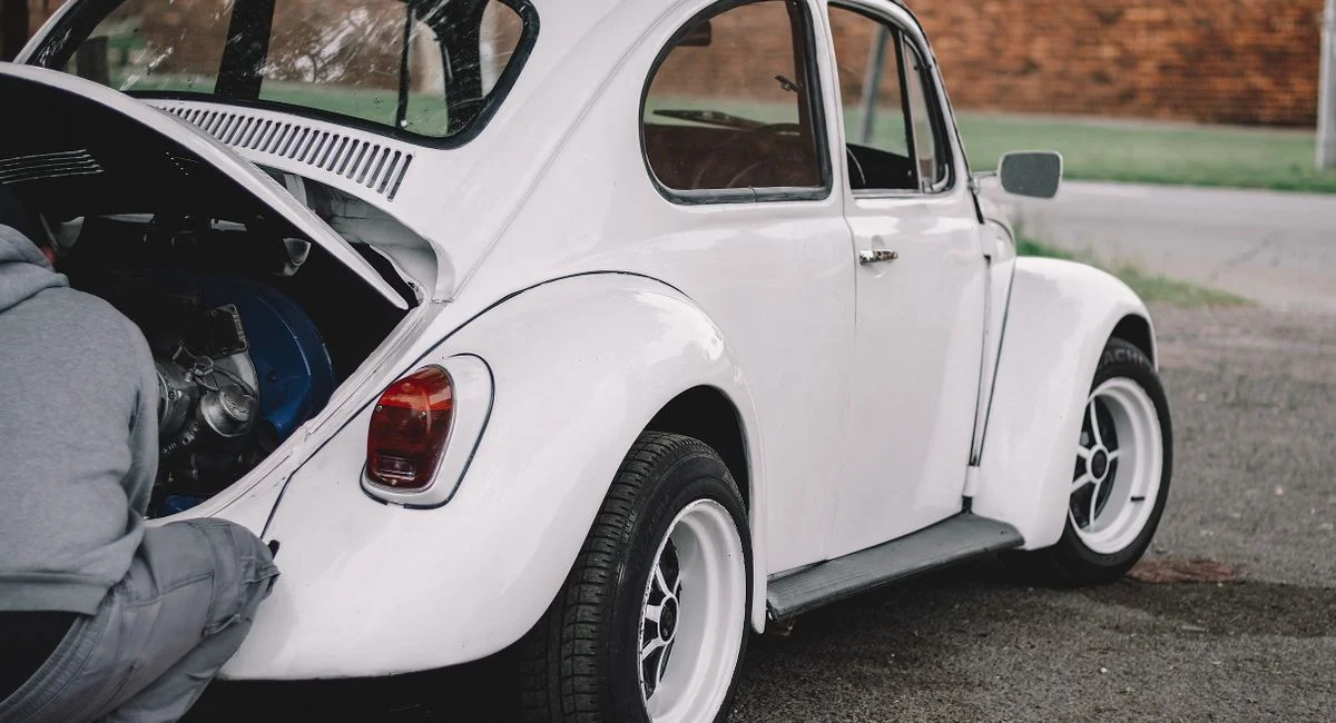 A mechanic working on a classic white Volkswagen Beetle engine outdoors.