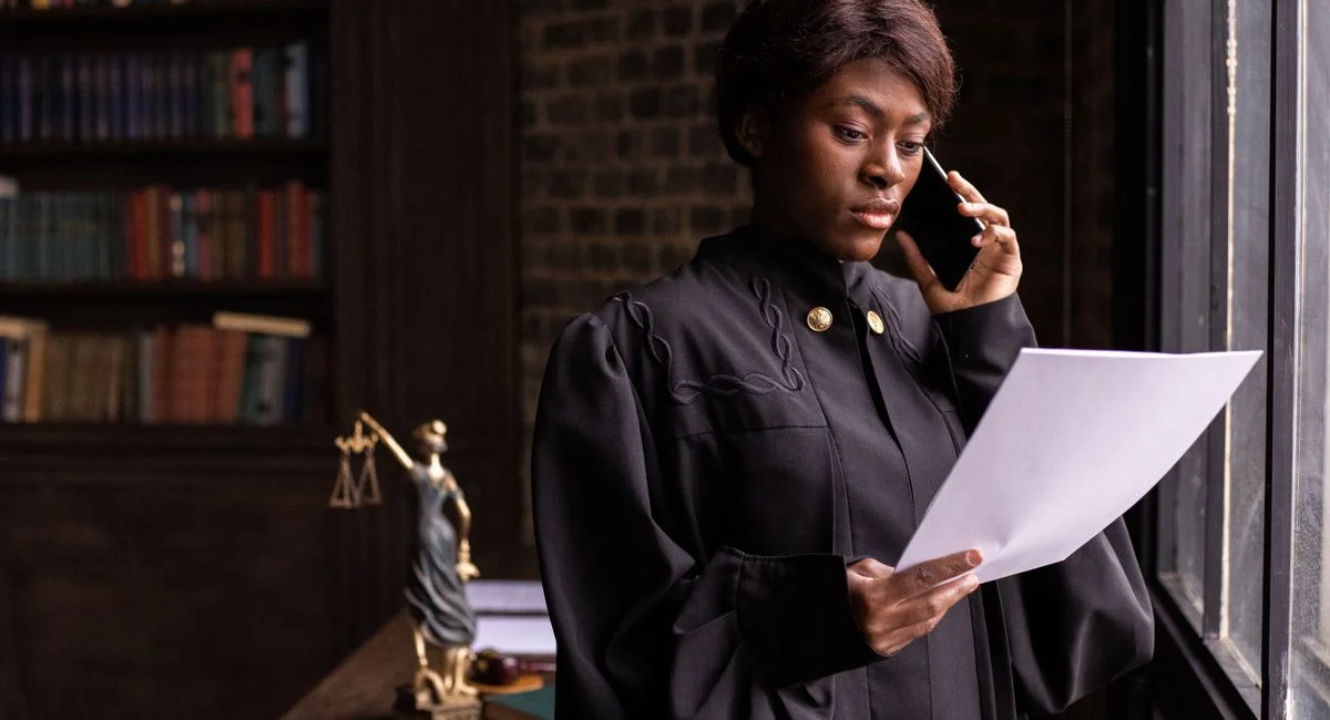 African American judge in robe, reading document and talking on phone by the office window.