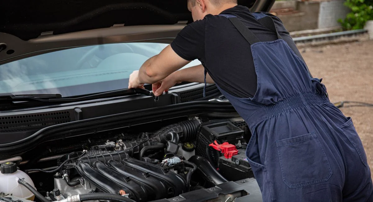 A mechanic in blue overalls works on a car engine outdoors, showcasing vehicle maintenance skills.