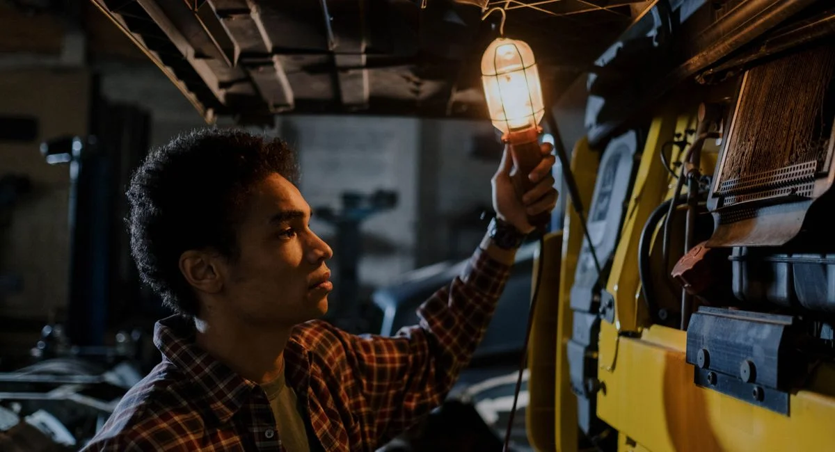 Auto mechanic inspecting a truck engine with a trouble light in a dimly lit garage.