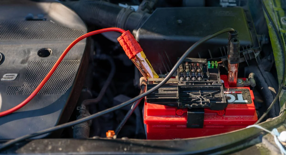 Close-up of a car battery with attached jumper cables in an engine bay.