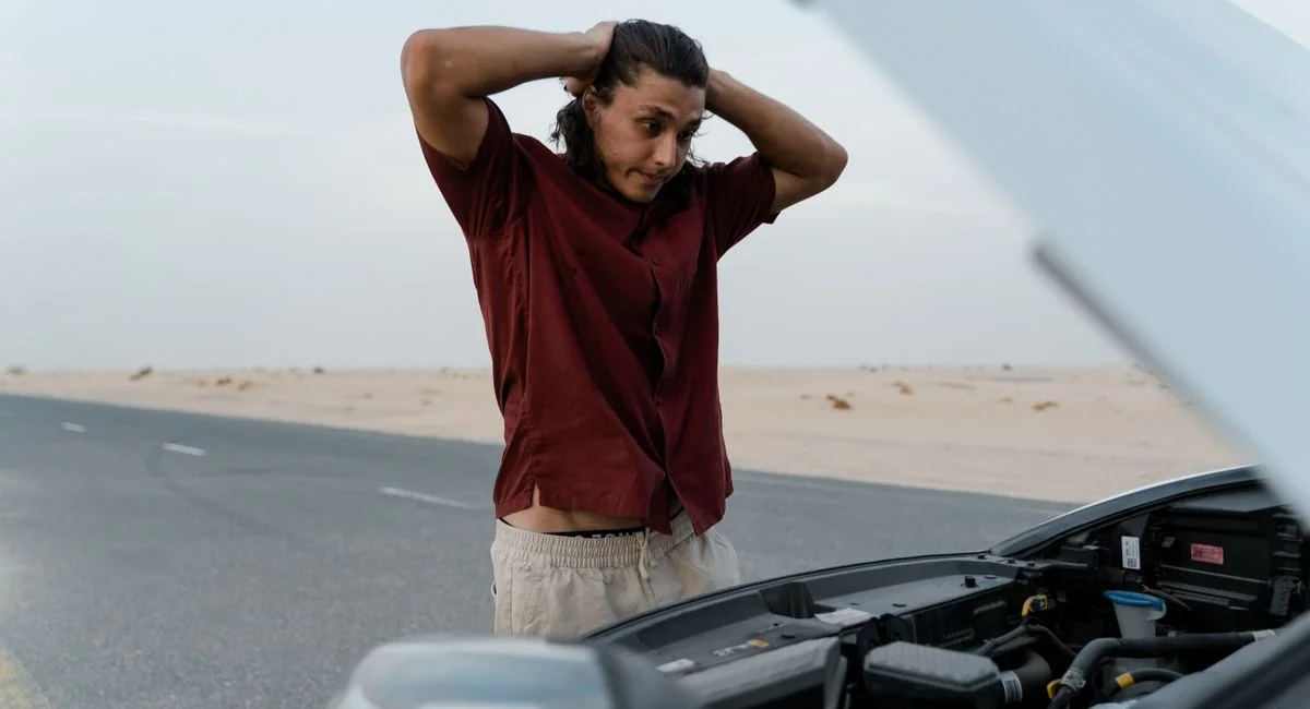 A frustrated man examining a broken car engine on a deserted road, showing stress and concern.