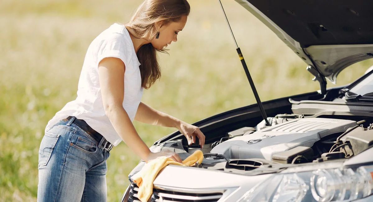 Side view of female driver in white t shirt and jeans checking damage of car engine and wiping details under open hood while 