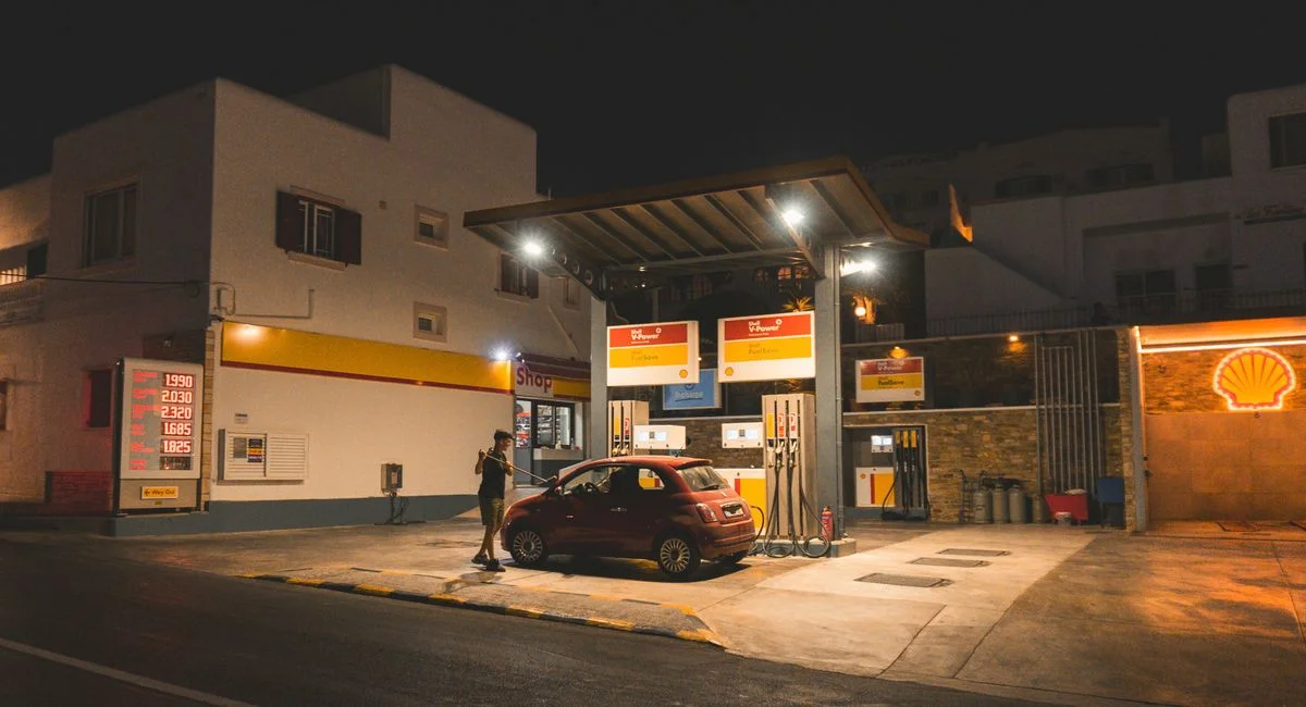 A person refuels a red car at a gas station at night, illuminated signs and lights.