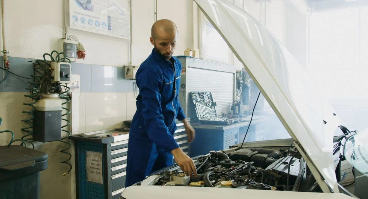 Mechanic in blue uniform inspecting a car engine in a workshop.