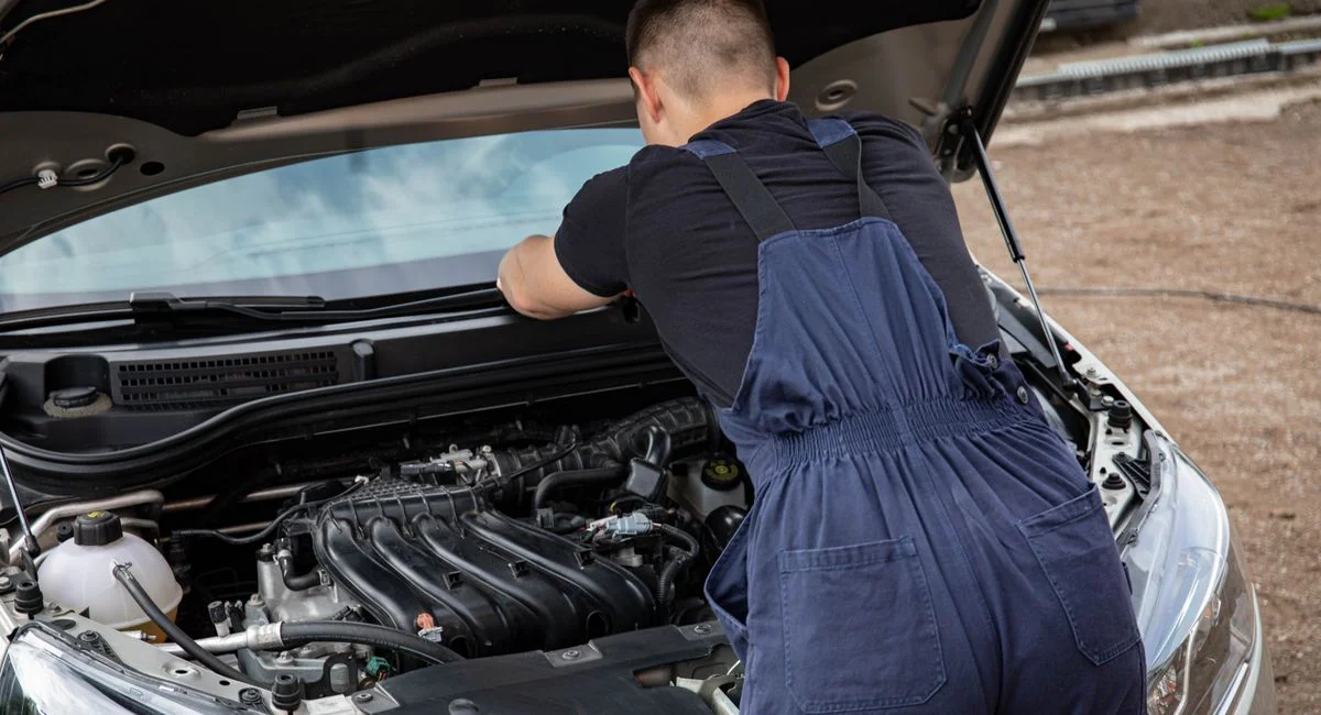 A mechanic in overalls fixing a car engine outdoors on a sunny day.