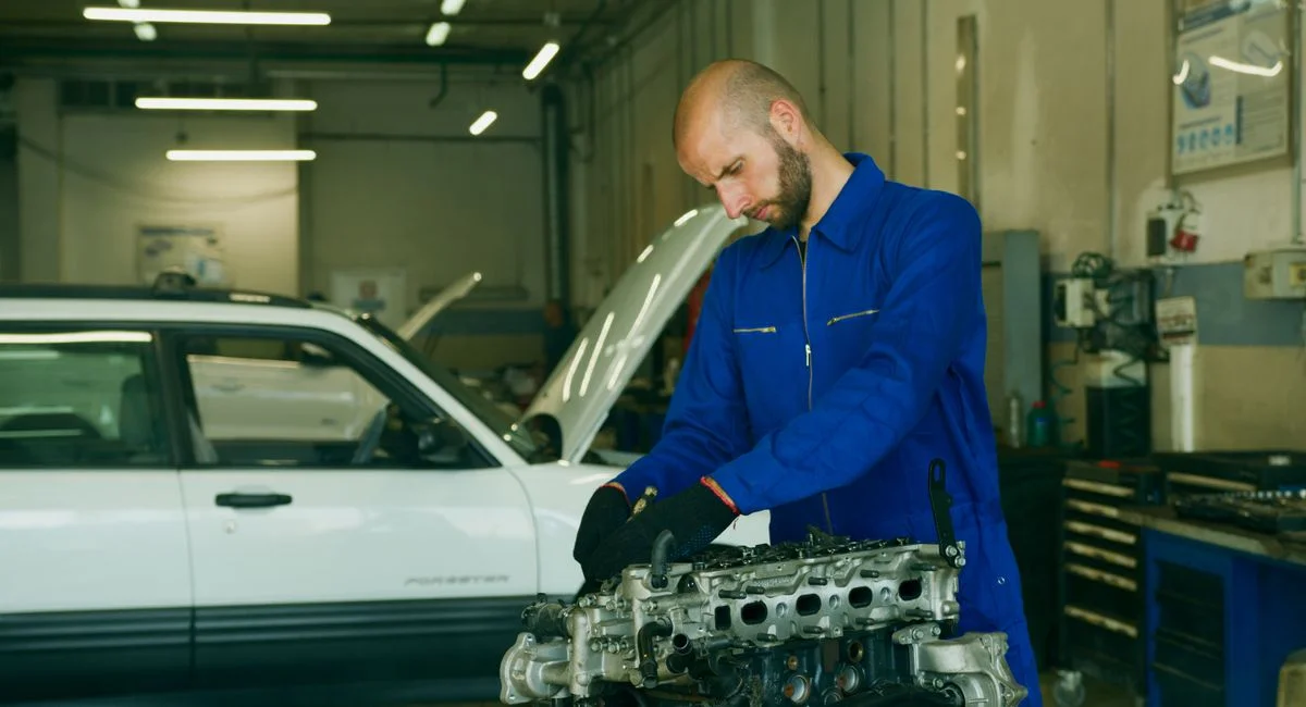 Mechanic in a blue coverall inspecting car engine in a garage workshop.