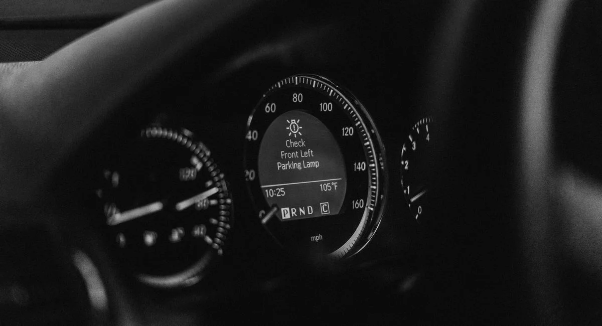 Close-up of a car dashboard in black and white with a parking lamp warning light.